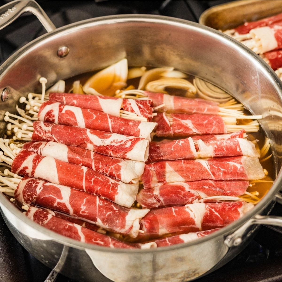 Raw beef enoki rolls arranged in a single layer over onions in simmering broth
