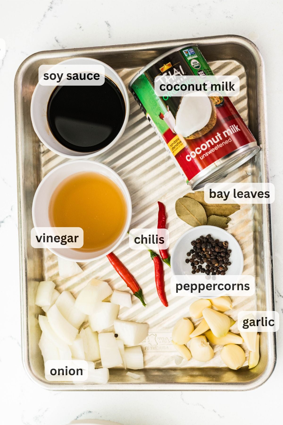 Labeled ingredients for chicken adobo sa gata on a metal tray: soy sauce, vinegar, coconut milk, bay leaves, chilies, peppercorns, garlic, and onion