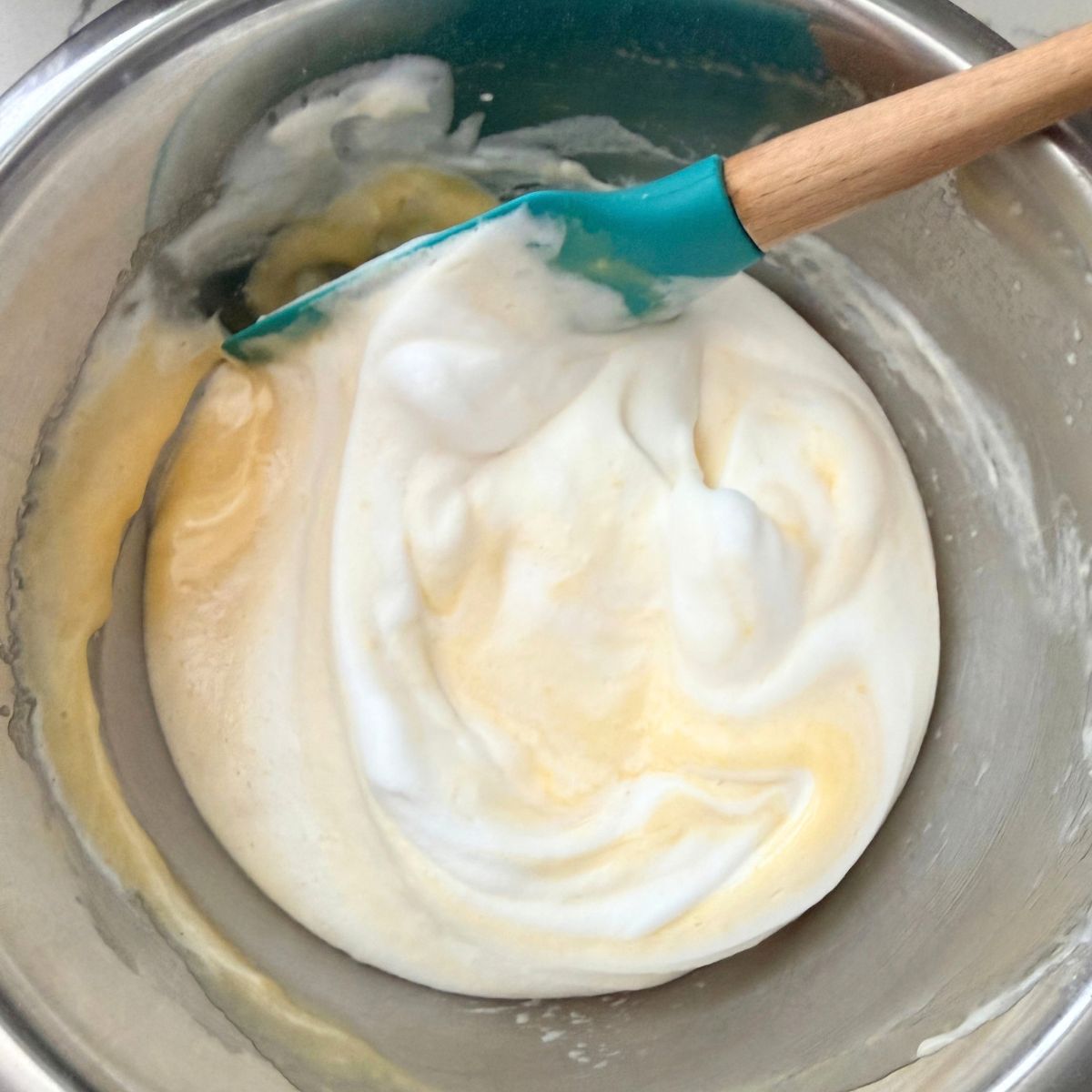 Glossy whipped egg whites being folded with a silicone spatula in a mixing bowl for yema cake.