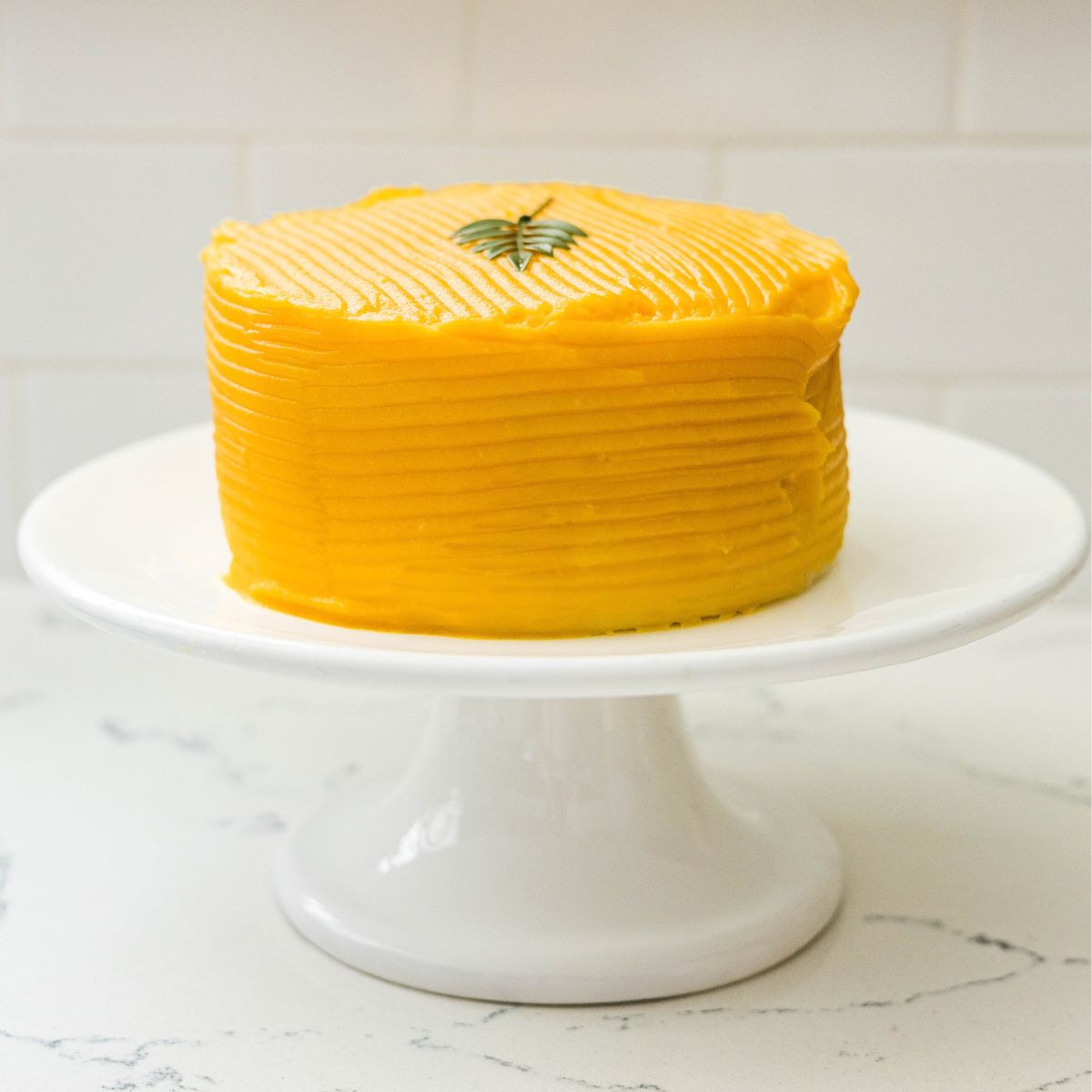 Yema cake with smooth golden frosting and a small green garnish on a white cake stand.