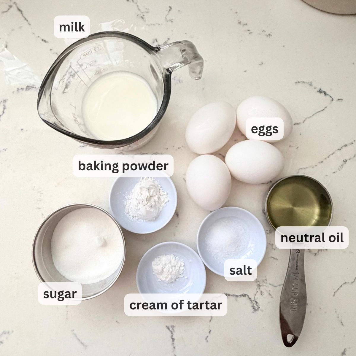 Overhead view of yema cake ingredients on marble, including milk, flour, baking powder, eggs, sugar, salt, and neutral oil.