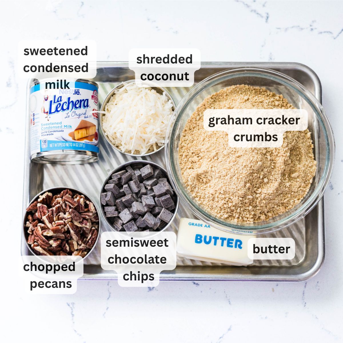 Ingredients for magic cookie bars: sweetened condensed milk, shredded coconut, graham cracker crumbs, semi-sweet chocolate chips, chopped pecans, and butter